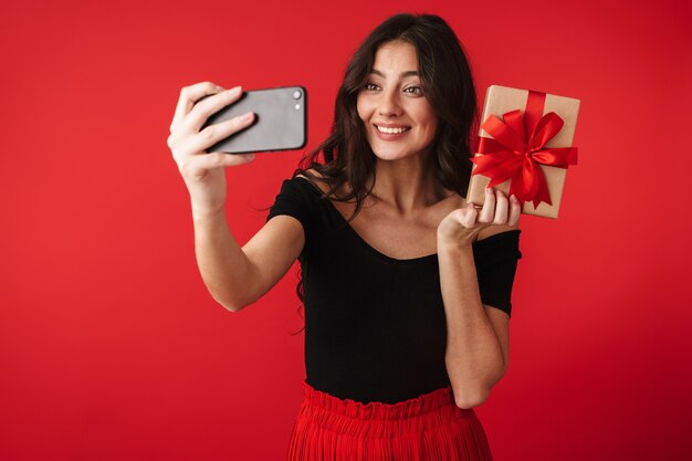 Счастливая молодая женщина в платье стоит изолированно над красным, держит подарочную коробку и делает селфи