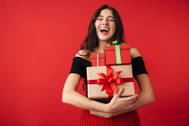 Happy young woman wearing a dress standing isolated over red, holding stack of gift boxes