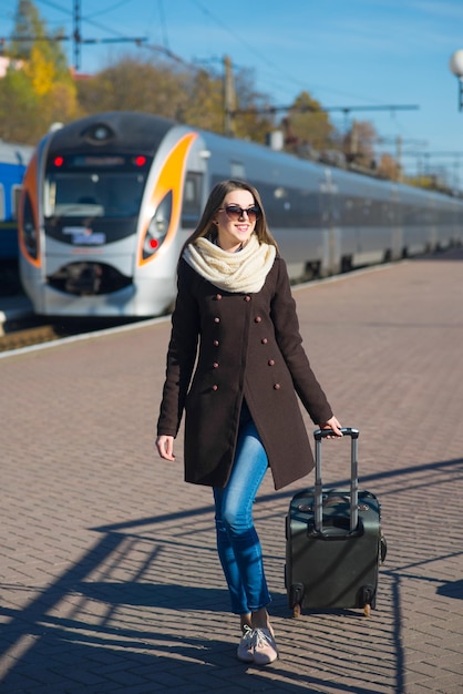 Felice giovane donna con gli occhiali vestita di cappotto con grande borsa da viaggio alla stazione ferroviaria sullo sfondo del treno