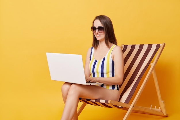 Happy young woman wearing striped onepiece swimsuit sitting on chair working on laptop pc isolated over yellow background doing her online work