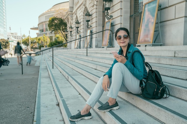 Happy young woman wearing sunglasses sitting on stairs and using her smartphone for communication via wifi internet outside san francisco city hall. smiling lady with backpack relax on street