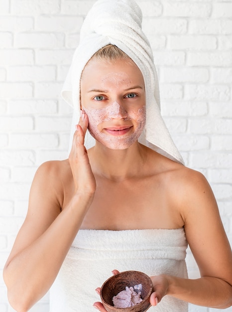 Happy young woman wearing white bath towels on head doing spa procedures applying facial scrub