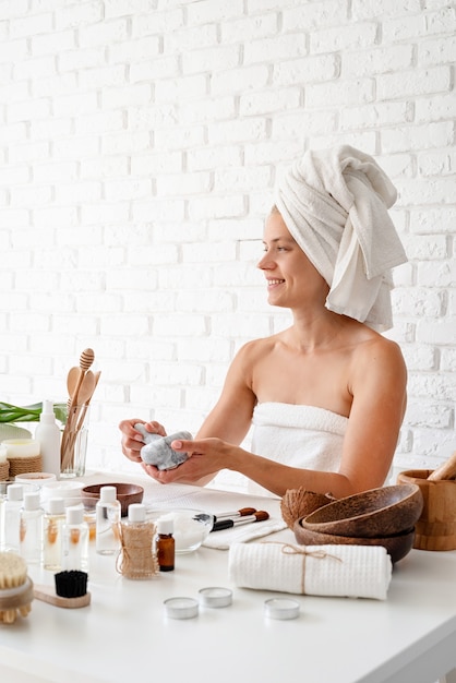 Happy young woman wearing white bathrobes towels on head doing spa procedures mixing natural ingredients in spa beauty salon