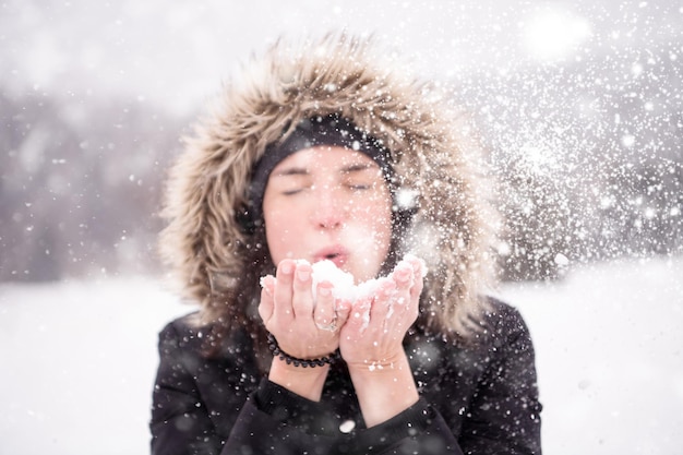 Happy young woman wearing winter clothes while blowing snow on snowy day with snowflakes around her in beautiful winter forest