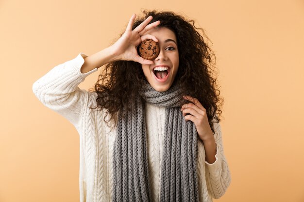 冬のスカーフを身に着けている幸せな若い女性がベージュの壁に孤立して立って、チョコレートクッキーを食べる