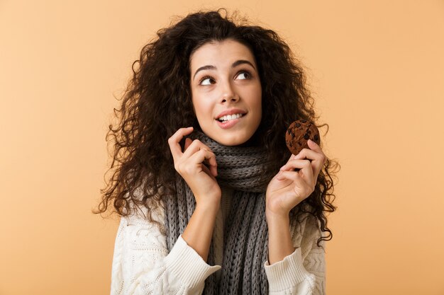 冬のスカーフを身に着けている幸せな若い女性がベージュの壁に孤立して立って、チョコレートクッキーを食べる