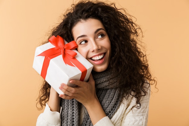 Happy young woman wearing winter scarf standing isolated over beige wall, holding present box