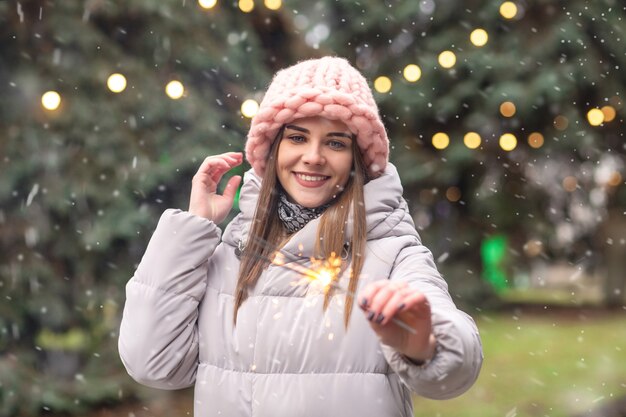 Happy young woman wears pink knitted hat and coat having fun with sparklers at the street near the Christmas tree during the snowfall