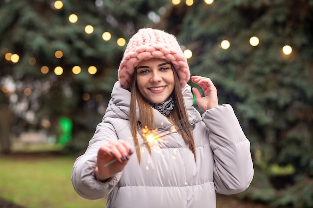 Happy young woman wears pink knitted hat and coat having fun with sparklers at the street near the Christmas tree