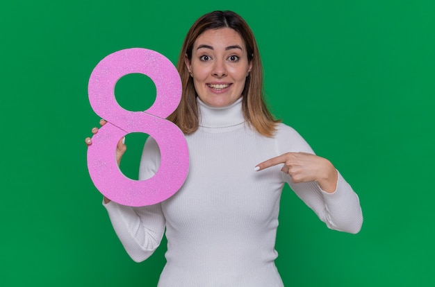 Foto felice giovane donna in bianco dolcevita tenendo il numero otto puntato con il dito indice a sorridere allegramente per celebrare la giornata internazionale della donna marzo