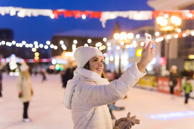 Felice giovane donna in inverno sulla pista di pattinaggio sul ghiaccio che scatta foto su smartphone selfie