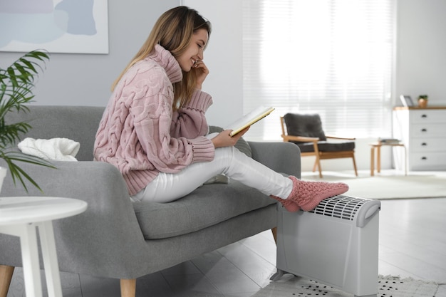 happy young woman with book warming feet on electric heater at home