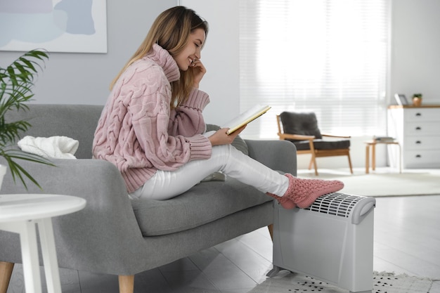 Happy young woman with book warming feet on electric heater at home