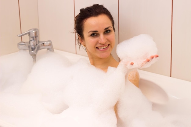 Happy young woman with bundle on her head lying in the tub full of foam in her bathroom