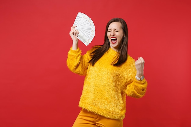 目を閉じて叫んで、勝者のジェスチャーをしている幸せな若い女性は、ドル紙幣でお金のファンを保持し、赤い背景で隔離の現金。人々の感情、ライフスタイルの概念。コピースペースをモックアップします。