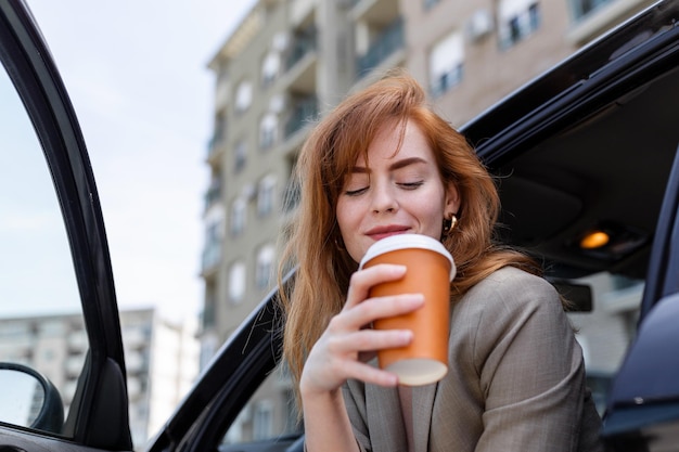 彼女の車にブレーキをかけているコーヒーと幸せな若い女性手に行くコーヒーと女性の側面図彼女の車でコーヒーを飲む若い女性