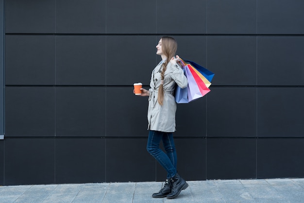Счастливая молодая женщина с красочными сумками и бумажным стаканчиком на улице. В