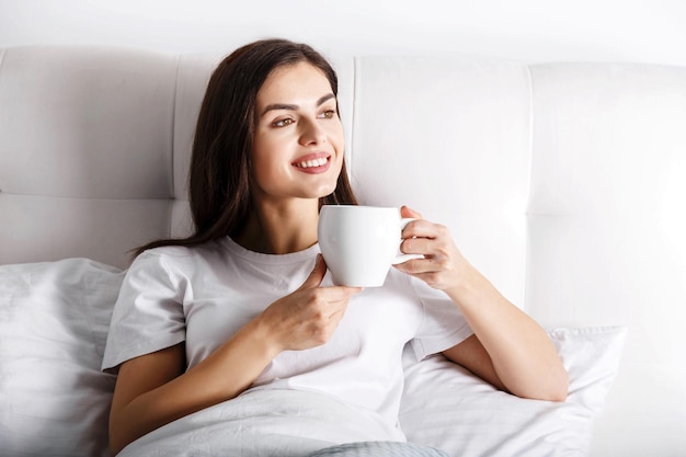 自宅の寝室のベッドでコーヒーやお茶と幸せな若い女性