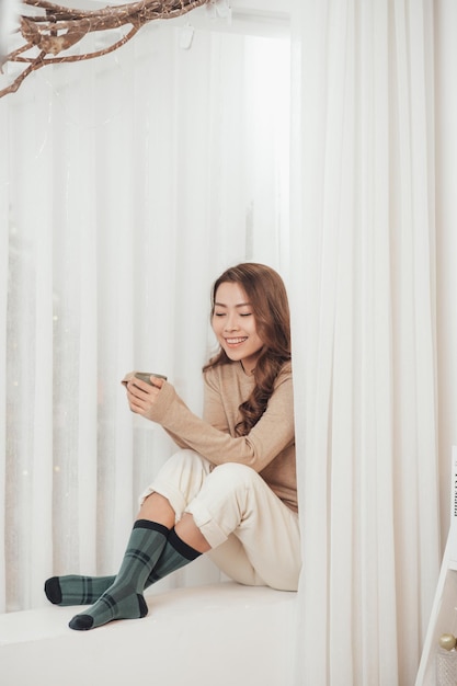 Happy young woman with cup of coffee or tea on the windowsill