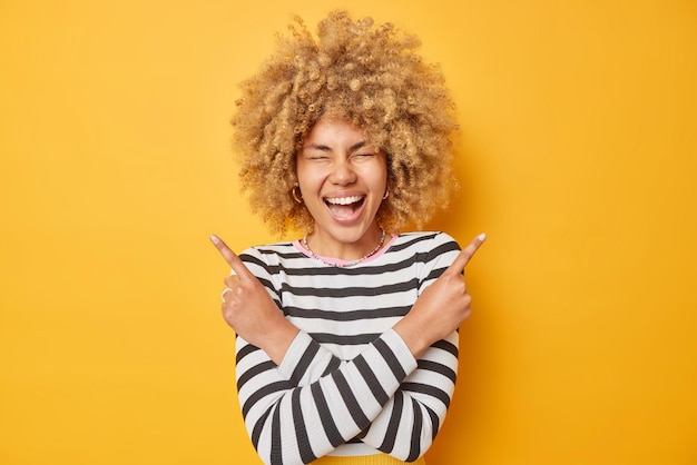 Happy young woman with curly blonde hair laughs out gladfully points sideways selects between two options picks two variants dressed in casual striped jumper isolated over yellow background