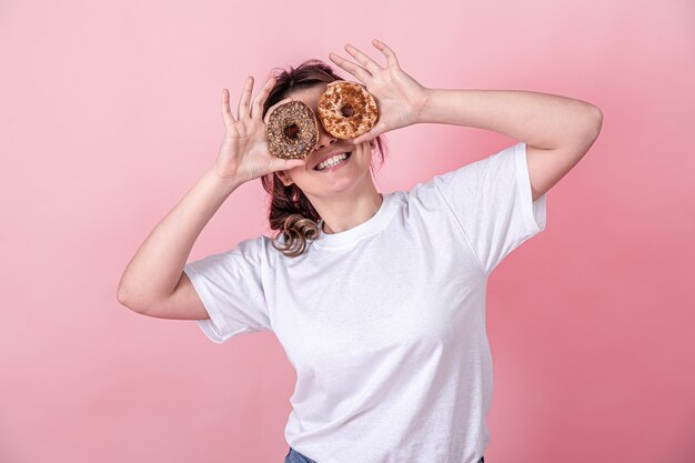 Happy young woman with donuts in her hands smile, holds pink donuts near her eyes her hands, pink background.