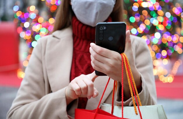 Una giovane donna felice con una maschera sul viso che porta delle borse della spesa in mano che compra con il suo smartphone