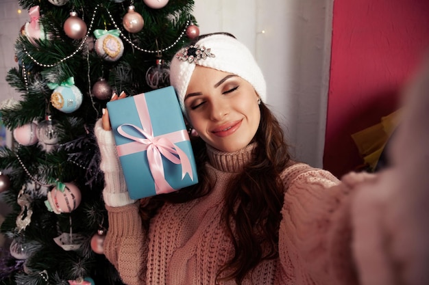 A happy young woman with a gift in her hand takes selfies against the Christmas tree Merry Christmas