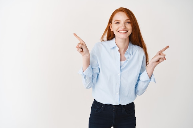 Happy young woman with ginger hair pointing fingers sideways, wearing office clothes and standing over white wall