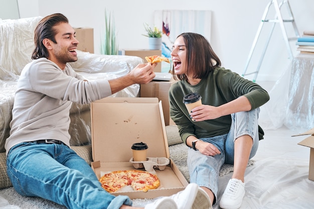 Felice giovane donna con un bicchiere di caffè da asporto aprendo la bocca mentre suo marito le dà una fetta di pizza durante il pranzo sul pavimento