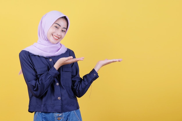 Una giovane donna felice con un'espressione facciale felice mostra qualcosa nella sua mano che indossa hijab e abiti casual in giallo