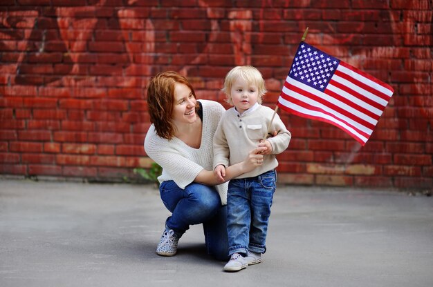 アメリカの国旗を保持している彼女の幼児の息子を持つ幸せな若い女。独立記念日のコンセプトです。