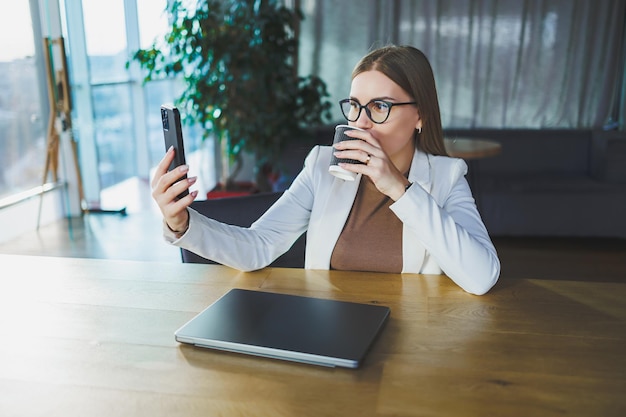 A happy young woman with long blond hair in stylish clothes is sitting at a table with a laptop enjoying a cup of fresh coffee and talking on the phone Work in a modern office with large windows