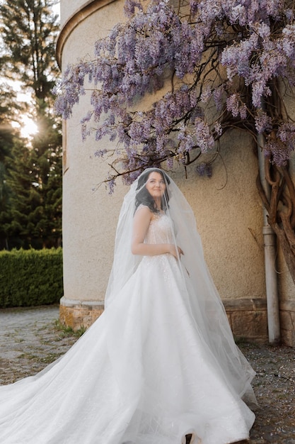 Happy young woman with long curly hair in a white dress under a veil near a garden