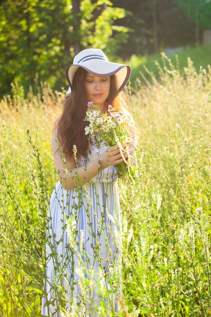 Happy young woman with long hair in hat and dress pulls her hands towards the plants while walking