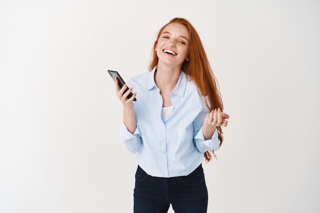 Happy young woman with long red hair and freckles laughing while using smartphone