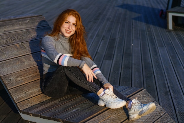 Happy young woman with long red hair is sitting in casual clothes in the park on a wooden deck on a sunny day and smiling