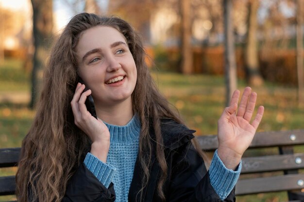 긴 물결 모양의 갈색 머리를 가진 행복한 젊은 여성이 공원 벤치에 앉아 전화 통화를 하고 가을 공원에서 몸짓 소녀를 합니다