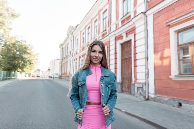 Happy young woman with smile with chic long blond hair in a summer sporty glamorous suit in a trendy blue denim jacket outdoors. Joyful European girl walks through the city. Modern street style.