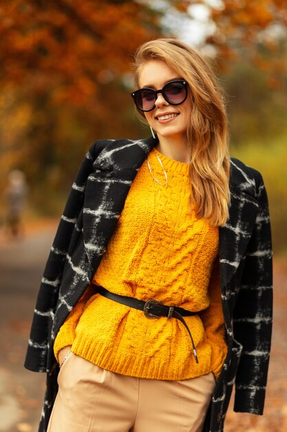 야외에서 황금빛 잎이 달린 노란색 니트 스웨터를 입은 세련된 코트에 선글라스를 끼고 웃는 행복한 젊은 여성