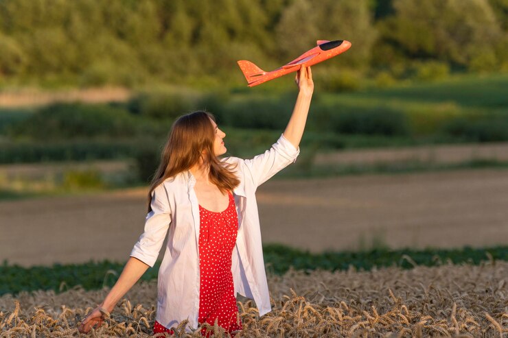 Happy young woman with toy airplane in hands in middle of wheat field in red dress and white shirt Concept of cheap air travel