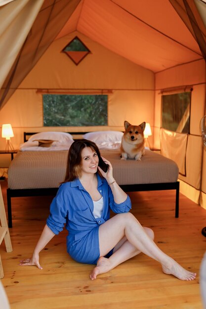 Happy young woman with Welsh Corgi Pembroke dog relaxing in glamping and talking phone Luxury camping tent for outdoor recreation and recreation Lifestyle concept