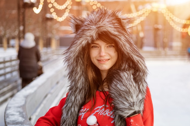 Happy young woman in a wolf hat in winter at the ice rink poses in a red sweater outside in the afternoon