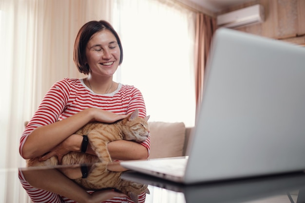 幸せな若い女性は、ラップトップとショウガの猫と一緒に家で働きます