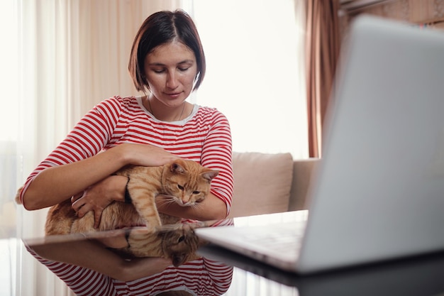 Happy young woman works at home with a laptop and a ginger
cat