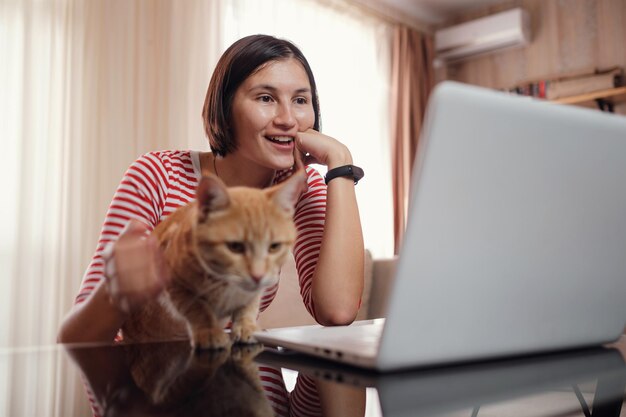 幸せな若い女性は、ラップトップとショウガの猫と一緒に家で働きます