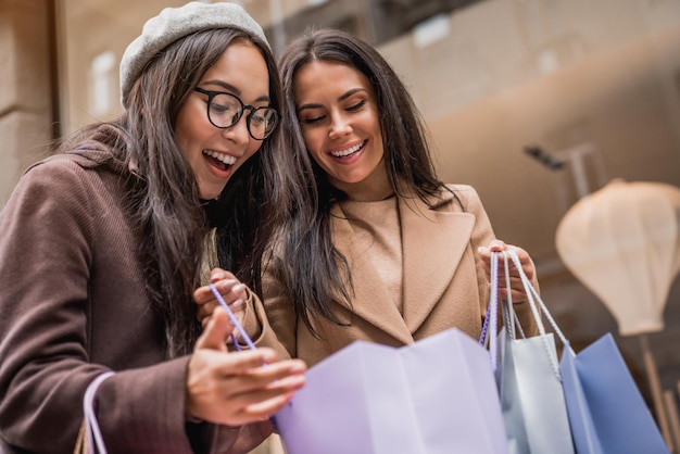 Photo happy young women looking into shopping bags of her friend near shop showcase outdoor