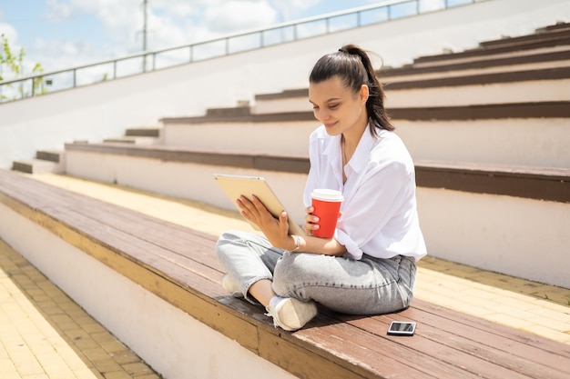 Giovani donne felici con la tazza di carta del caffè della tenuta della compressa che gode del giorno soleggiato che si siede in anfiteatro.