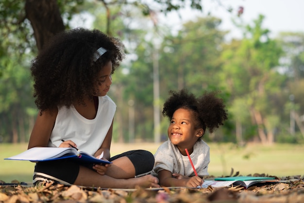 Happy younger sister smiling and looking at her older sister while lying drawing in the coloring book for kids in the park.Family and relationship concept.