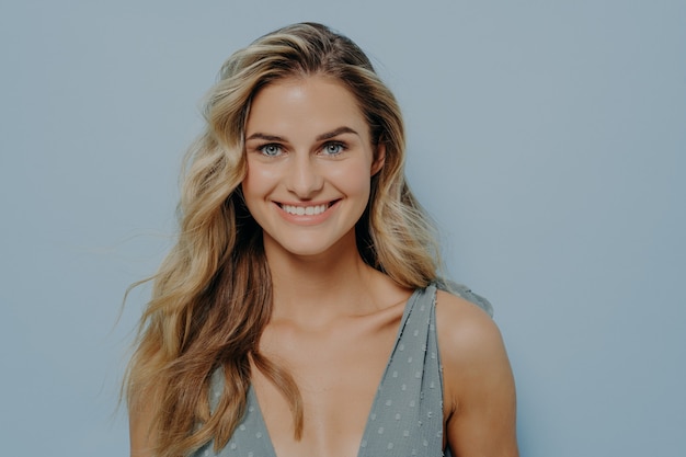 Happy youthful blonde girl in blue dress smiling with bright and cheerful smile, showing teeth, looking confident and positive while standing isolated over blue background in studio. Happiness concept