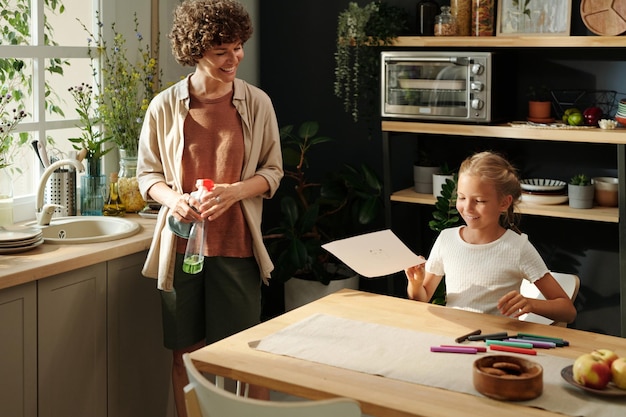 Happy youthful girl showing her drawing to mother with plastic bottle